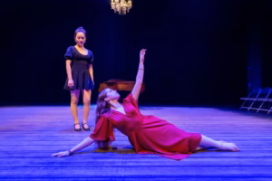 Sarah Genne as Linda Lee Thomas in Interlochen Arts Academy's Musical Theatre Composer's Showcase: Cole Porter, reclining on stage in a red gown with one arm raised, as another performer in black looks on.
