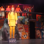 Sarah Genne as Dr. Jane Borden Osgood in Stagedoor Manor's production of Anyone Can Whistle, speaking atop a wooden soap crate with the ensemble cast behind her.