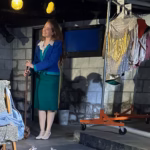 Sarah Genne as Mallory in Stagedoor Manor's production of According to the Chorus, smiling backstage in a blue and teal costume, holding a wooden pole attached to a wooden dog puppet.